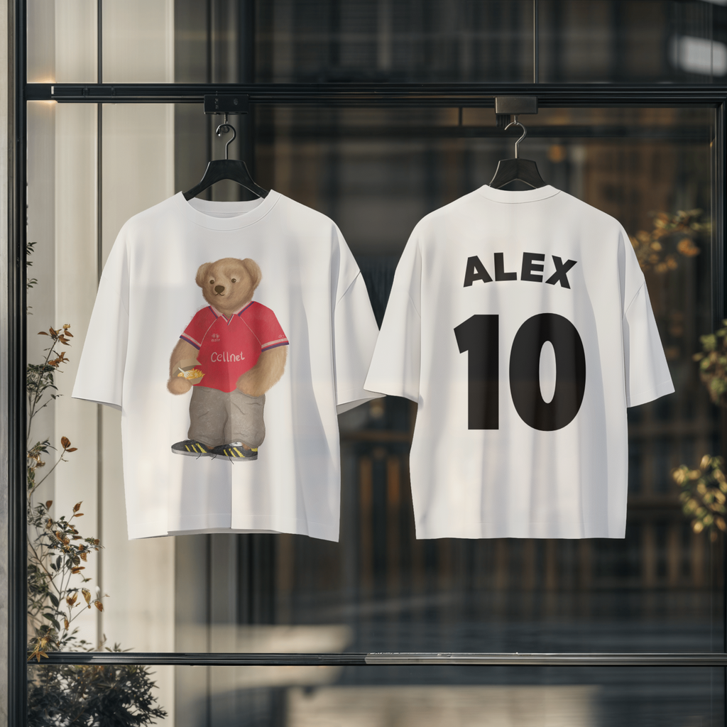 Two white t-shirts with a teddy bear graphic and text on hangers against a blurred indoor background.