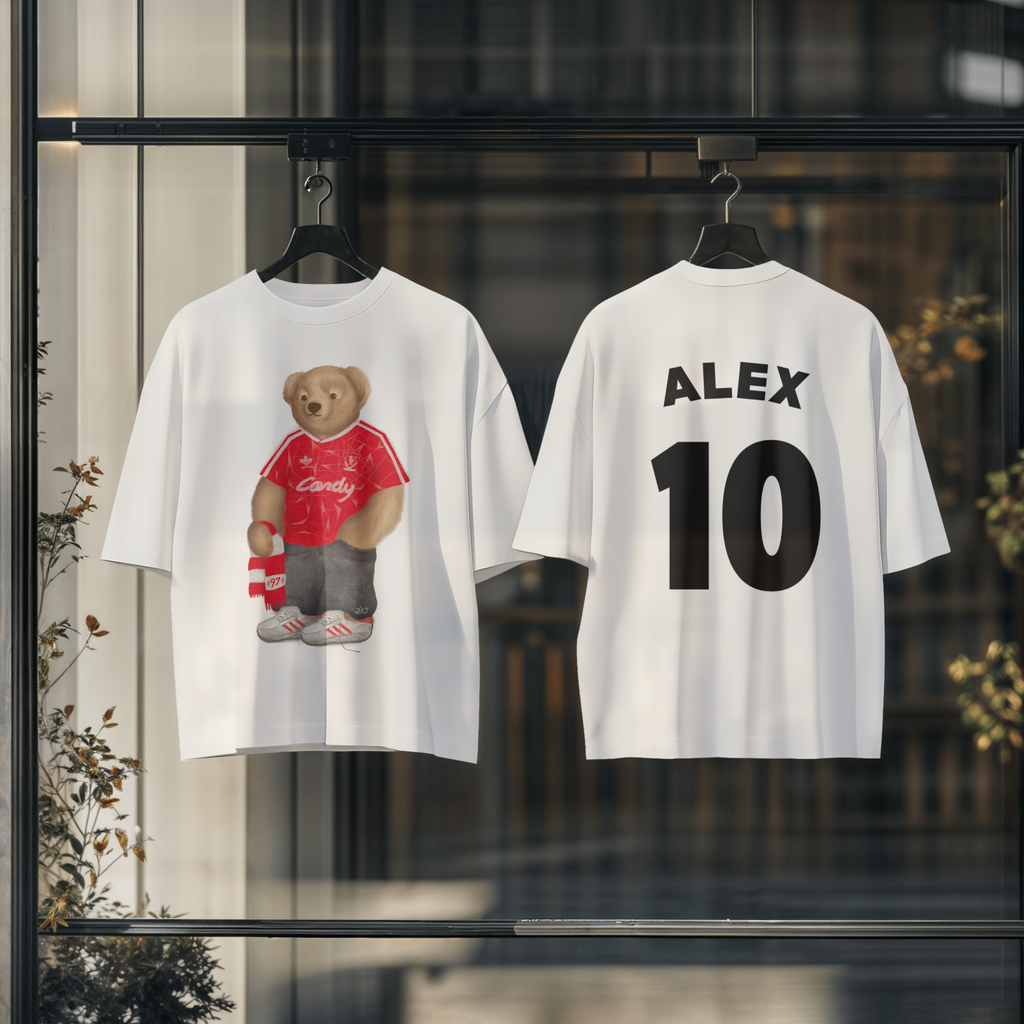 Two white t-shirts with a teddy bear graphic and text on hangers against a blurred indoor background.