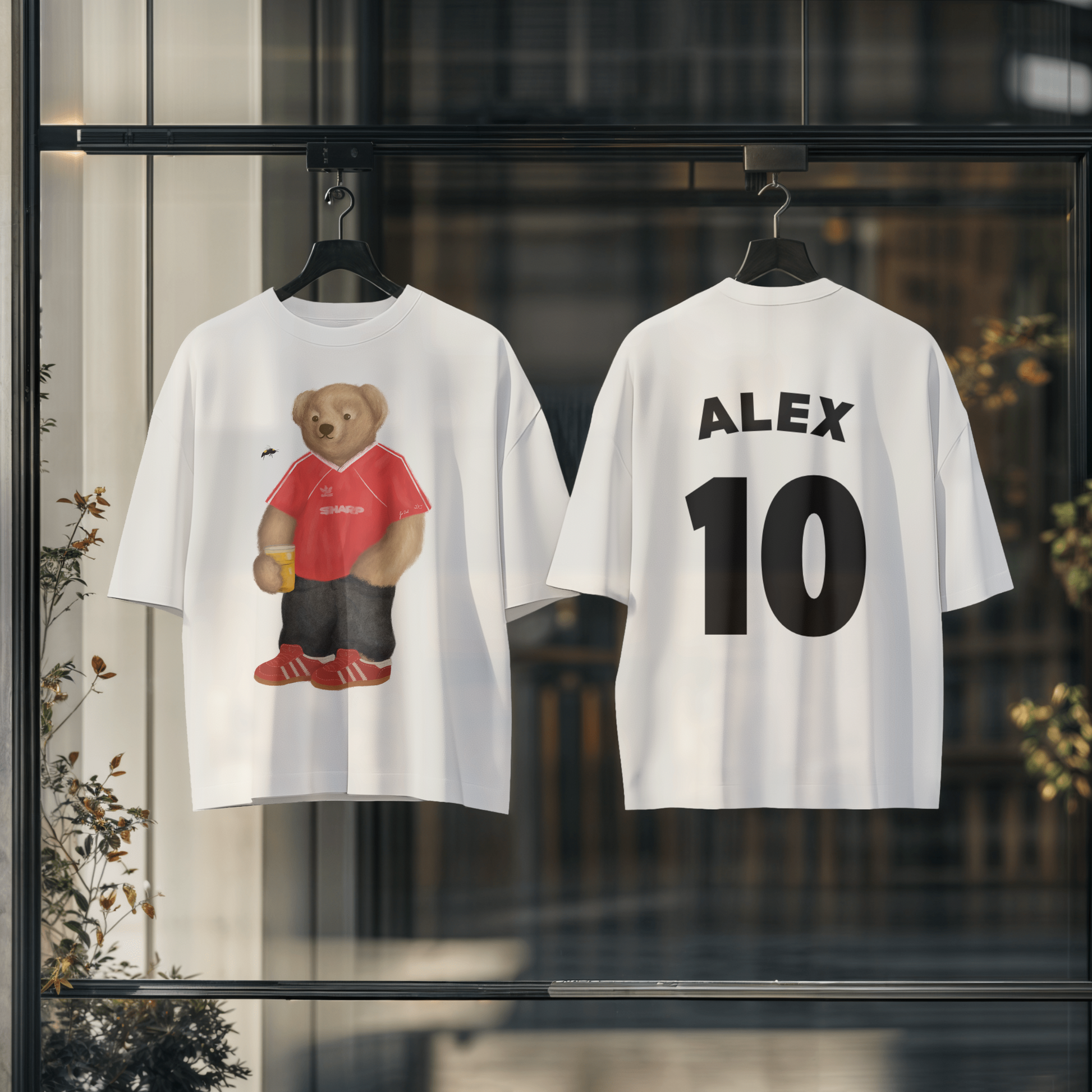 Two white t-shirts with a bear graphic and text on hangers against a blurred indoor background.