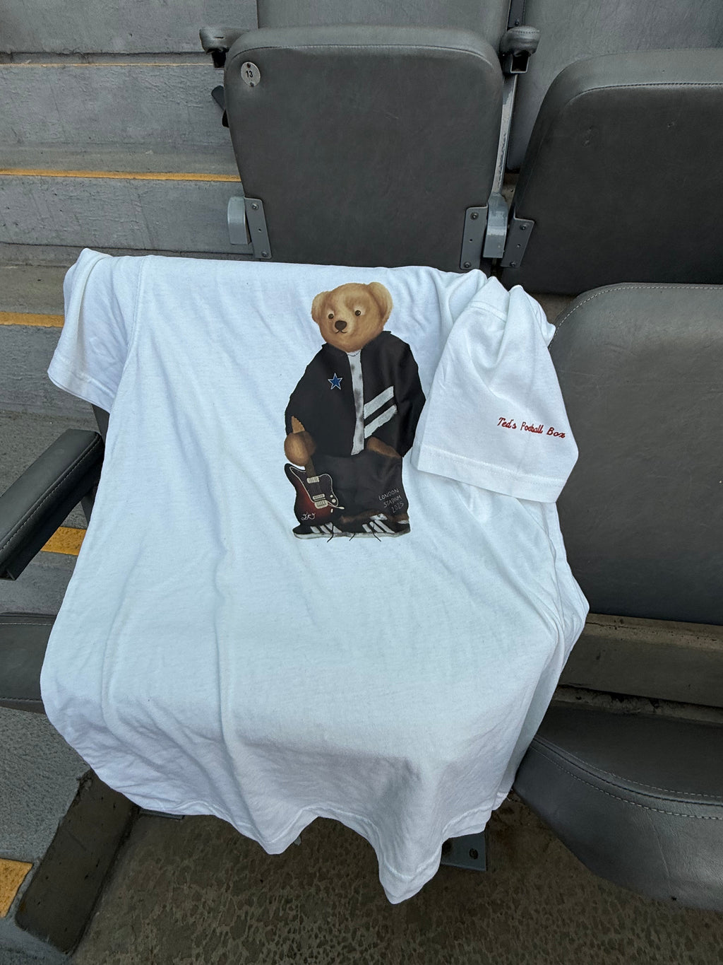 White t-shirt with a graphic of a bear wearing a jacket on a chair.