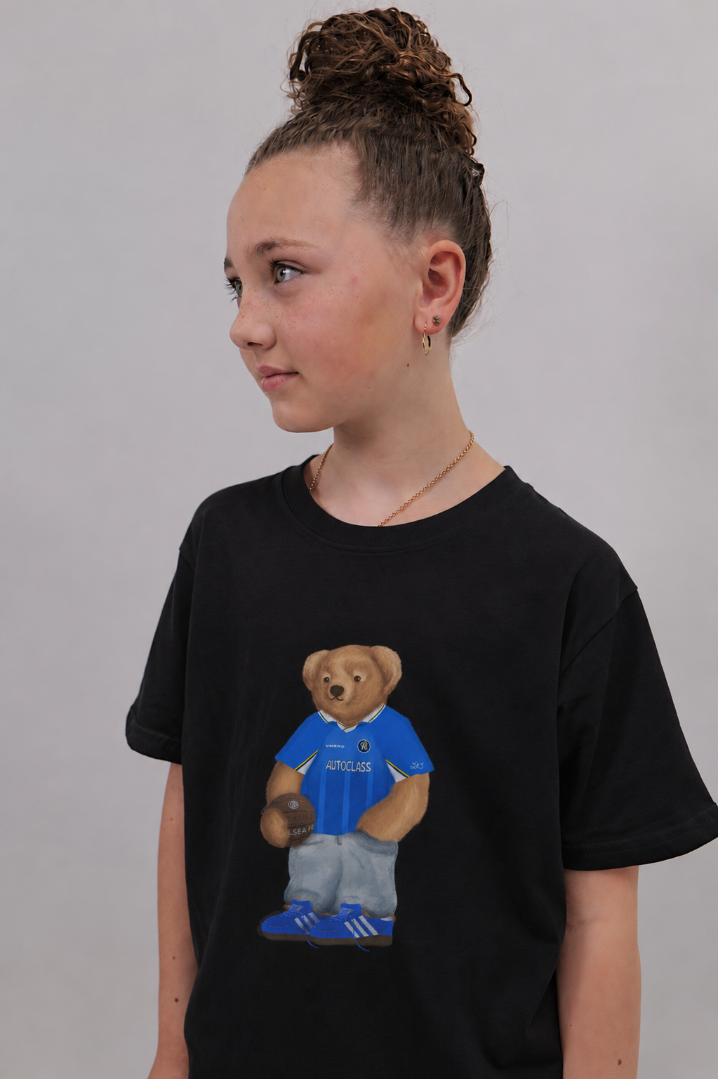 A child wearing a black short-sleeve t-shirt with a graphic of a teddy bear in a blue and gray soccer jersey. The child is shown in profile against a plain light gray background.