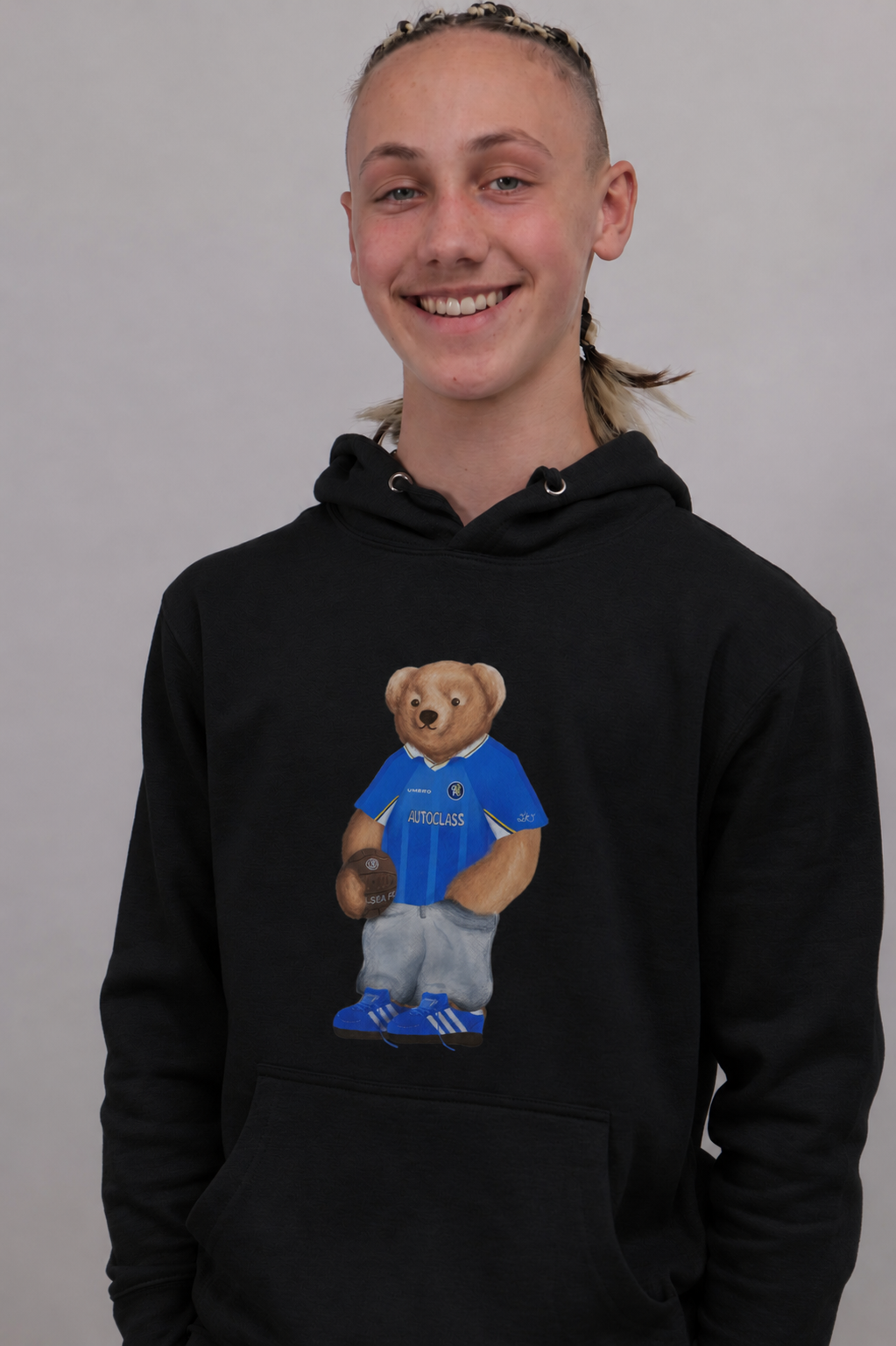 A smiling young person wearing a black pullover hoodie with a graphic of a teddy bear in a blue soccer jersey on the front, photographed against a plain light gray background.