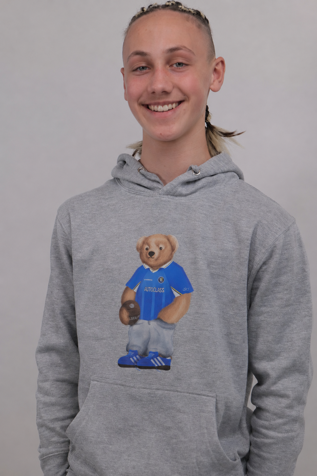 A smiling young person wearing a heather gray pullover hoodie with a teddy bear graphic on the chest, standing against a plain light gray background.