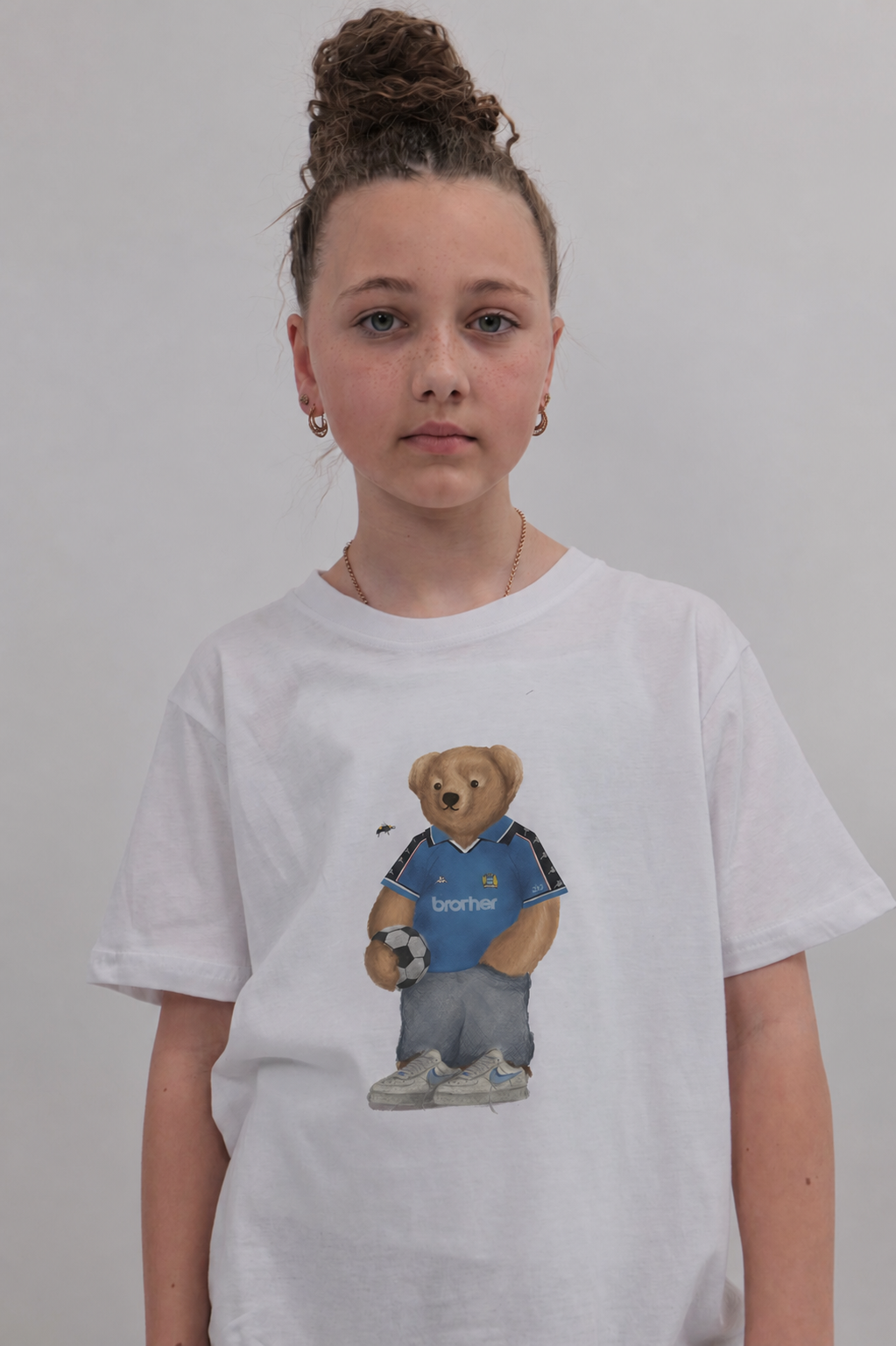 A young girl with her hair in a bun wears a white t-shirt featuring a graphic of a teddy bear in a blue soccer jersey holding a soccer ball. The background is a plain light gray wall.