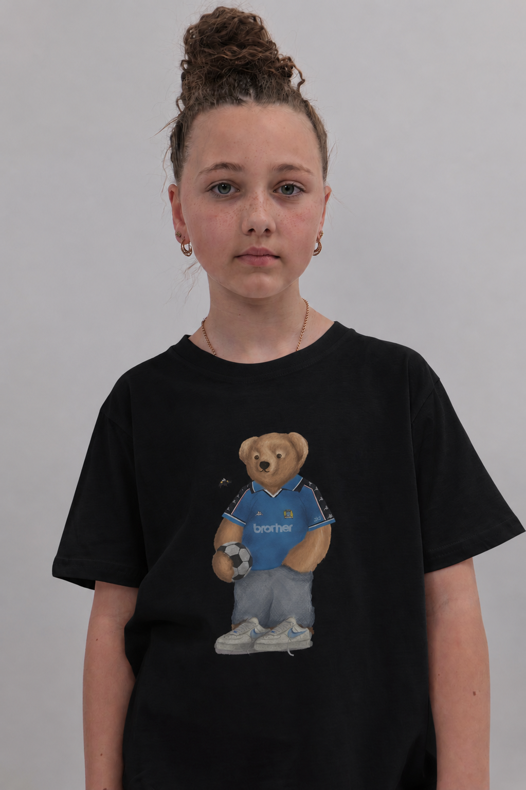 A young girl with her hair in a high bun wearing a black t-shirt with a graphic of a teddy bear in a blue soccer jersey. The background is a plain light gray wall.