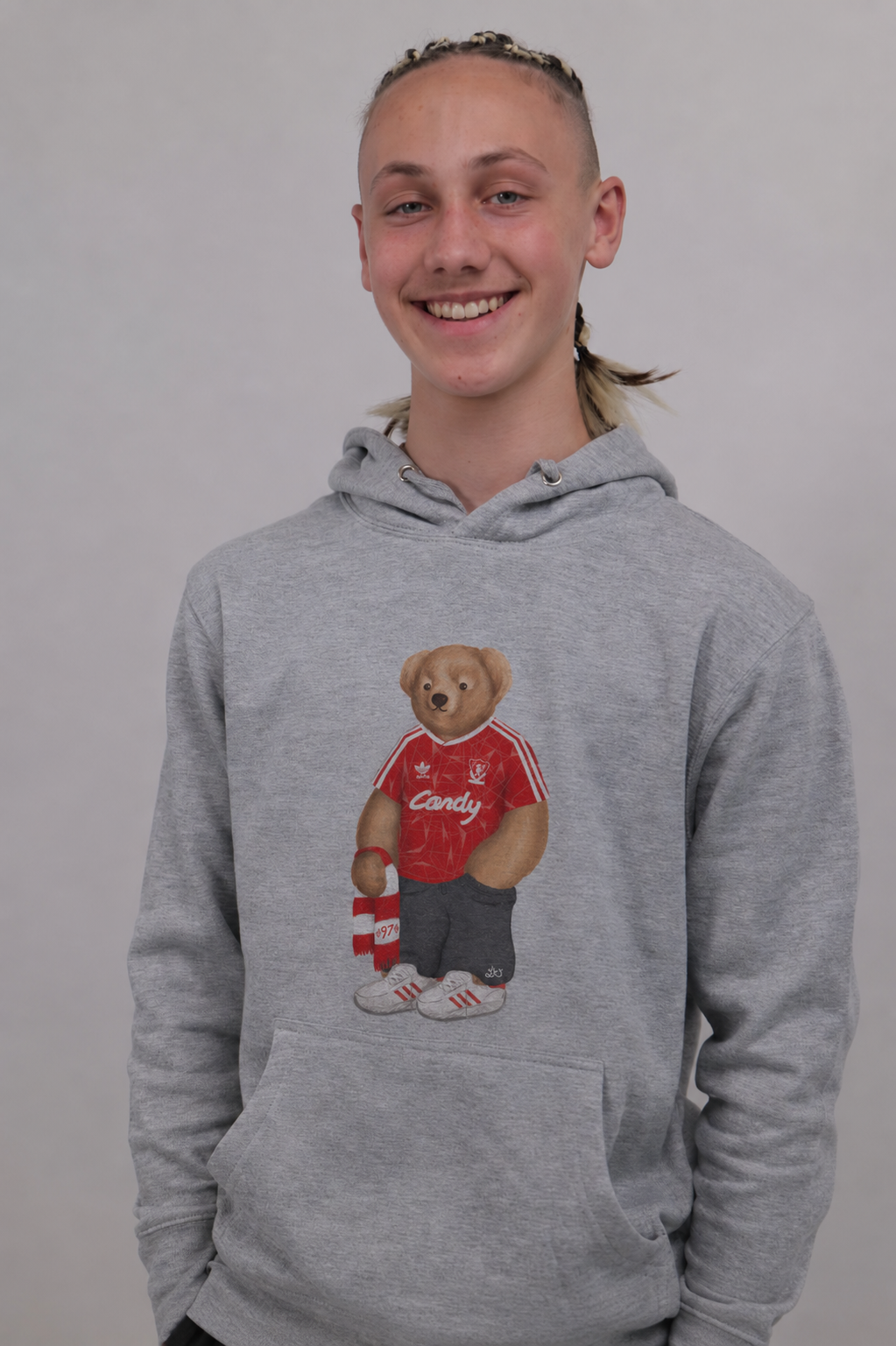 A smiling young person wearing a heather gray pullover hoodie with a teddy bear graphic on the chest, standing against a plain light gray background.