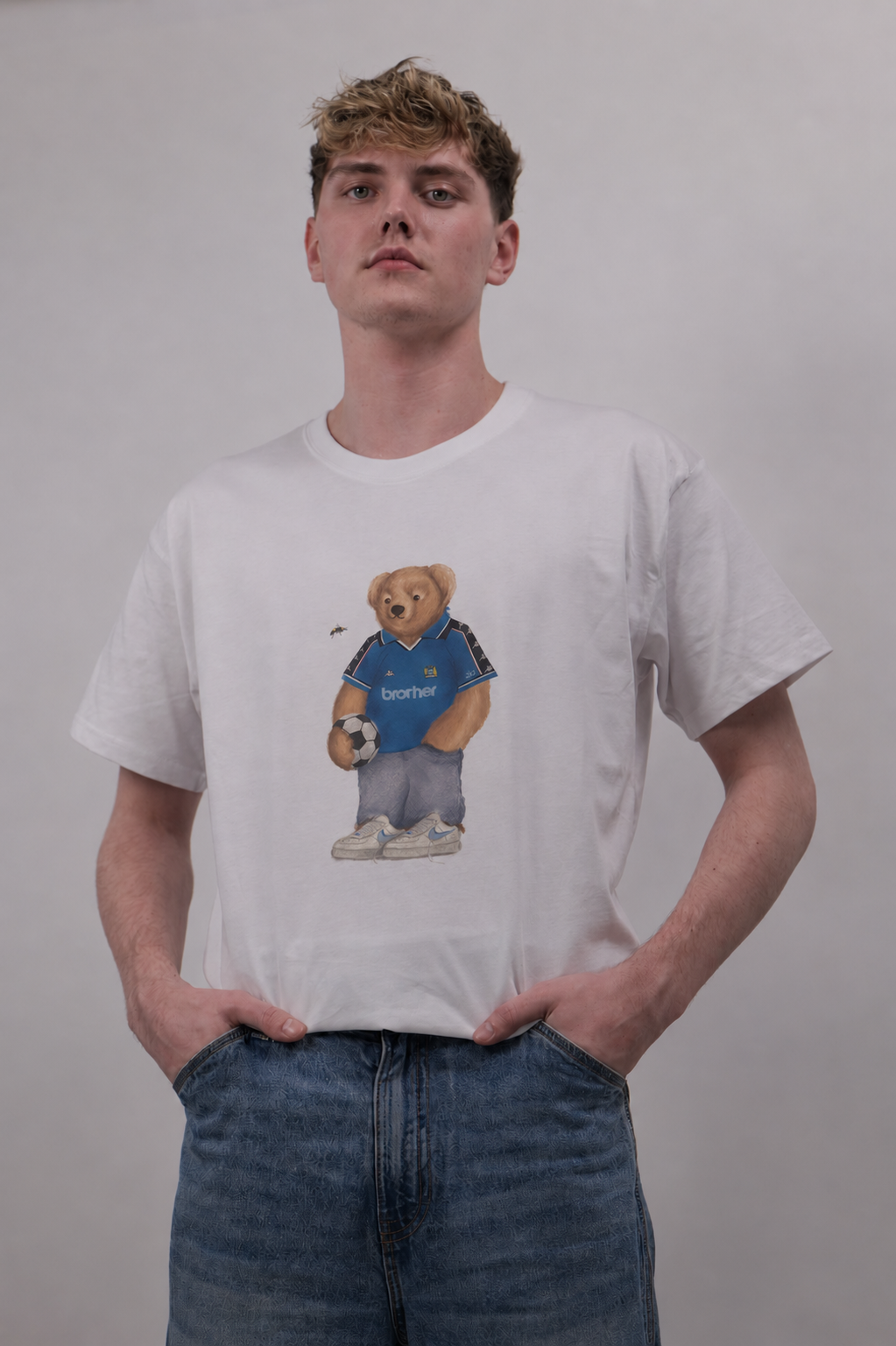 A young man wearing a white crewneck t-shirt with a graphic of a teddy bear in a blue soccer jersey, paired with blue jeans, against a plain light gray background.