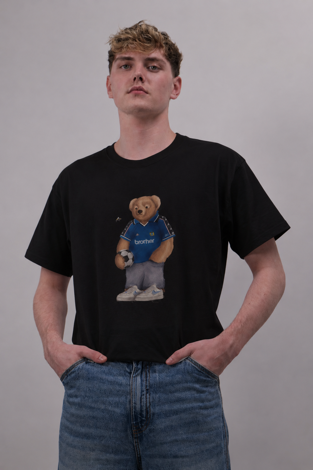 A young man wearing a black crewneck t-shirt with a graphic of a teddy bear in a blue soccer jersey holding a soccer ball. The t-shirt features the word 'Sportful' on the chest.