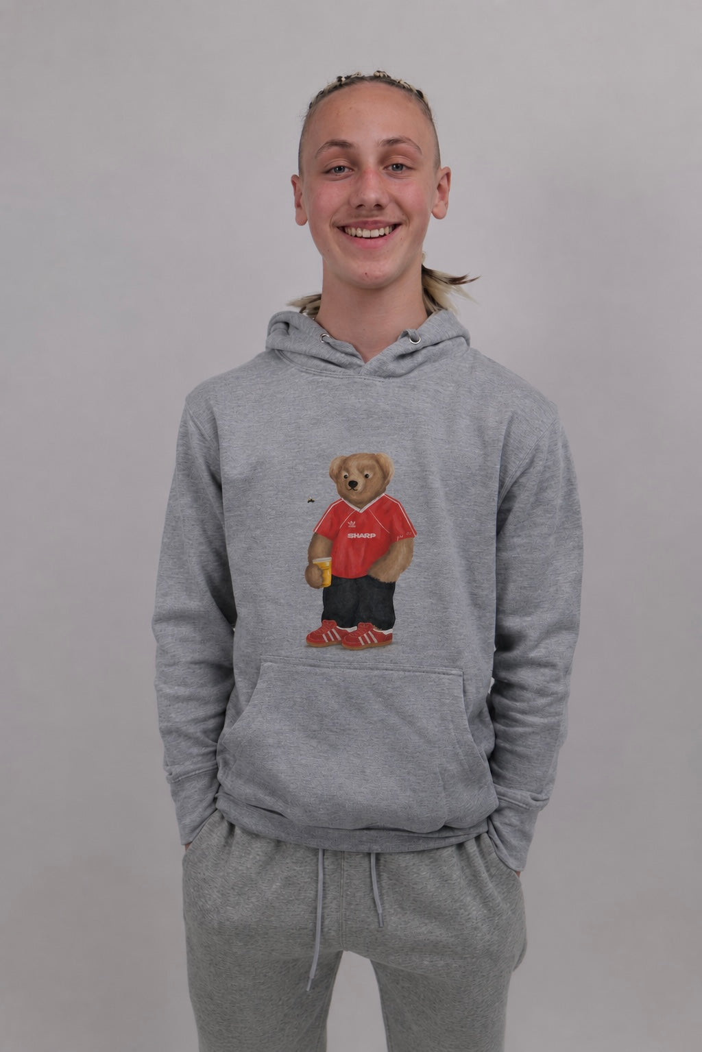 A smiling young person wearing a heather gray pullover hoodie with a teddy bear graphic on the front, paired with matching gray sweatpants, against a plain light gray background.