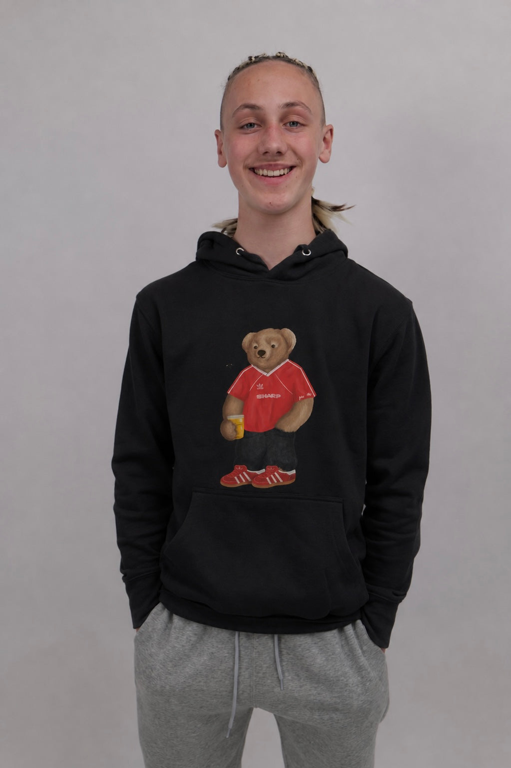 A smiling young man wearing a black pullover hoodie with a teddy bear graphic holding a red drink, paired with gray sweatpants, against a plain light gray background.