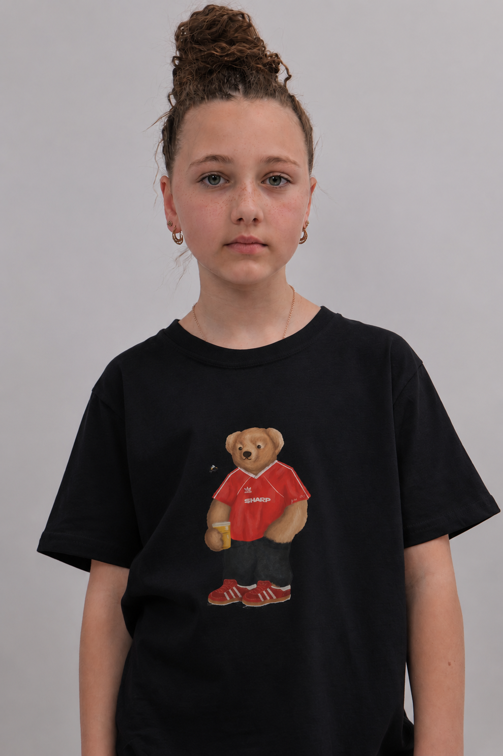 A young girl with her hair in a high bun wears a black t-shirt featuring a graphic of a teddy bear in a red shirt and red sneakers. The background is a solid light gray studio wall.