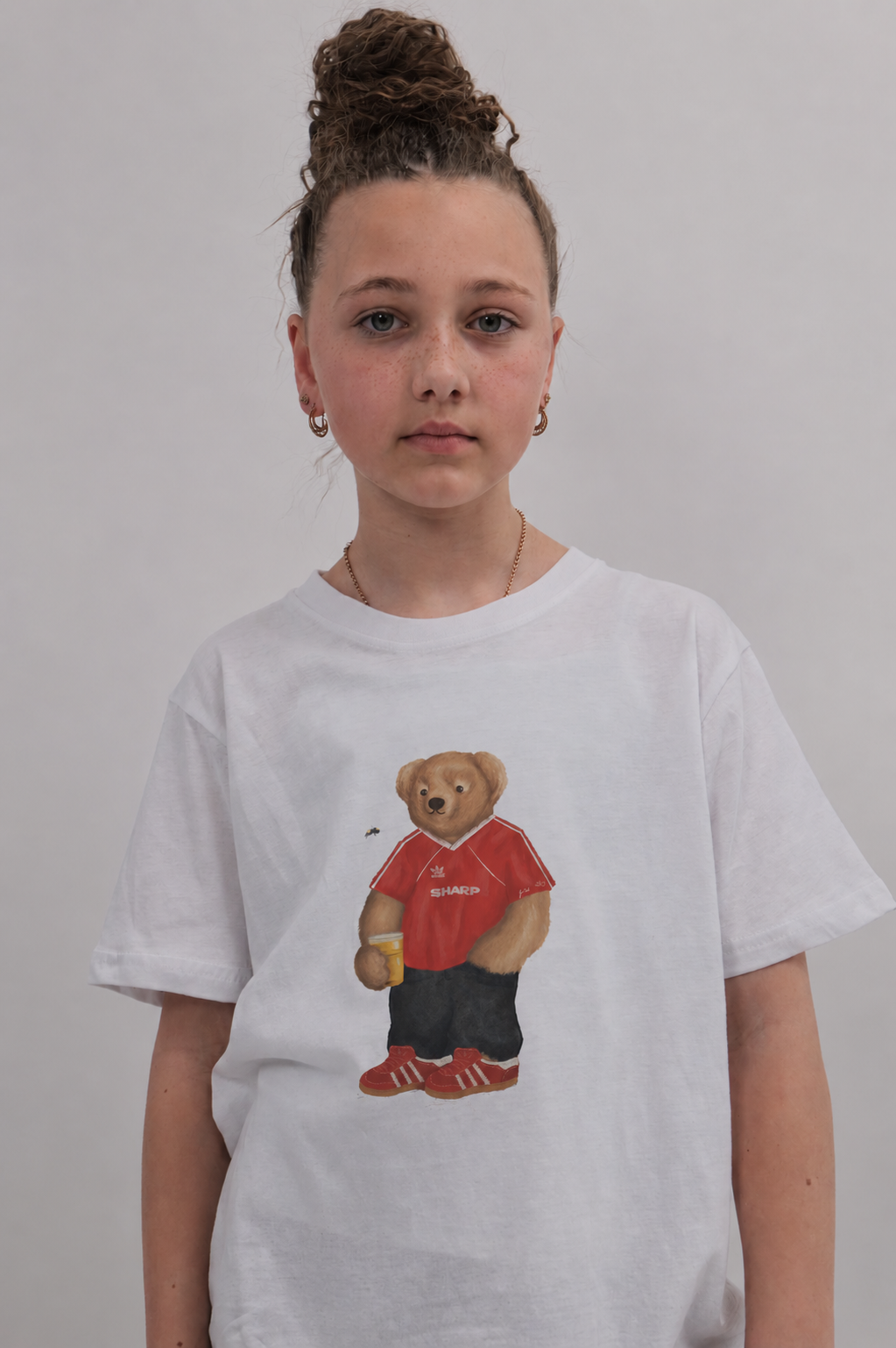 A young girl with her hair in a high bun wears a white crewneck t-shirt featuring a graphic of a teddy bear in a red shirt and black shorts, set against a plain light gray background.