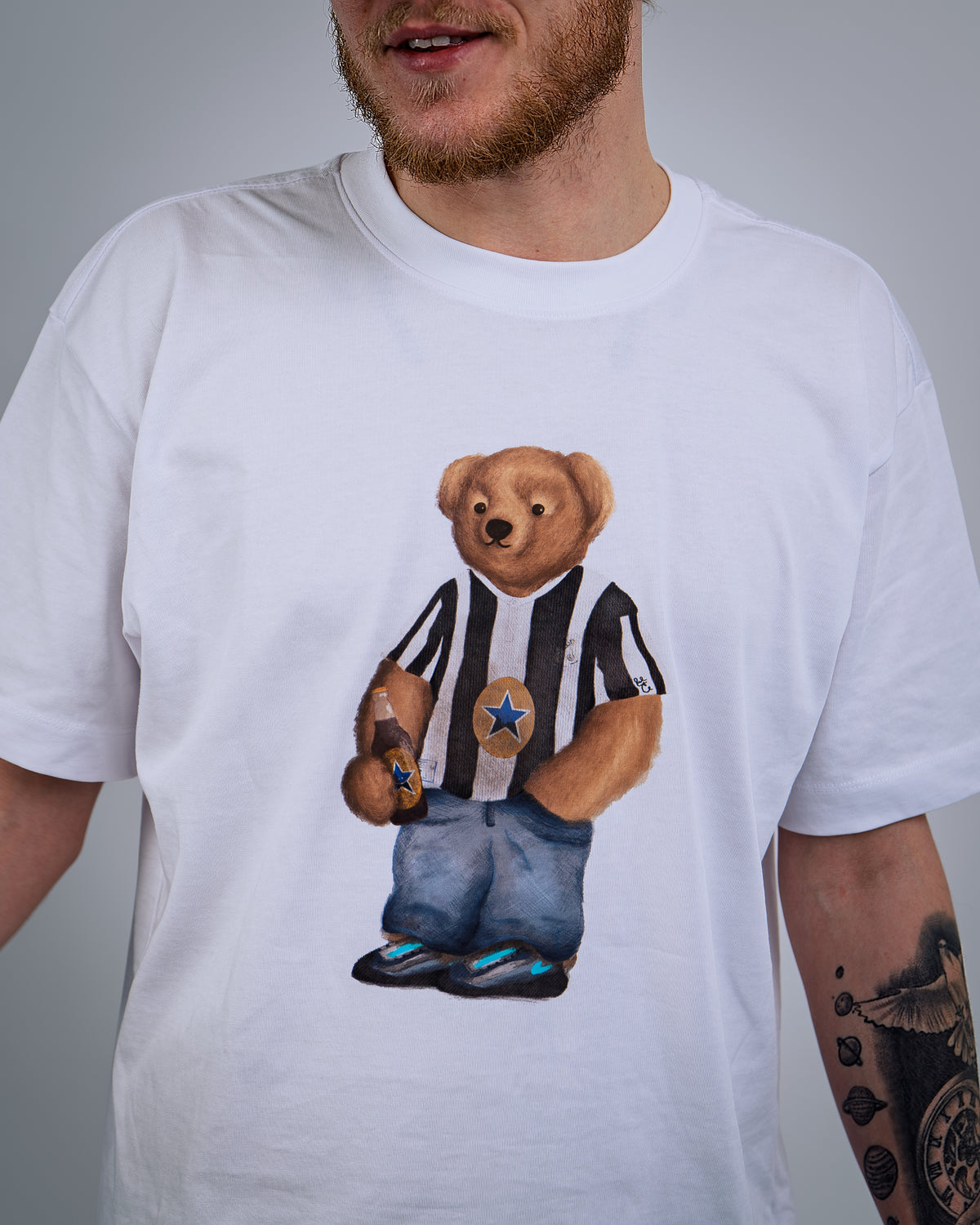 White t-shirt with a teddy bear graphic wearing a striped shirt and jeans on a gray background.
