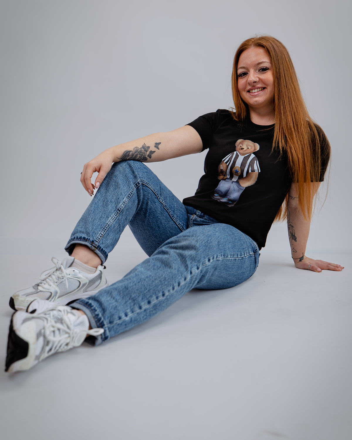 Woman sitting on a light gray background wearing a black t-shirt with a graphic design, blue jeans, and white sneakers.