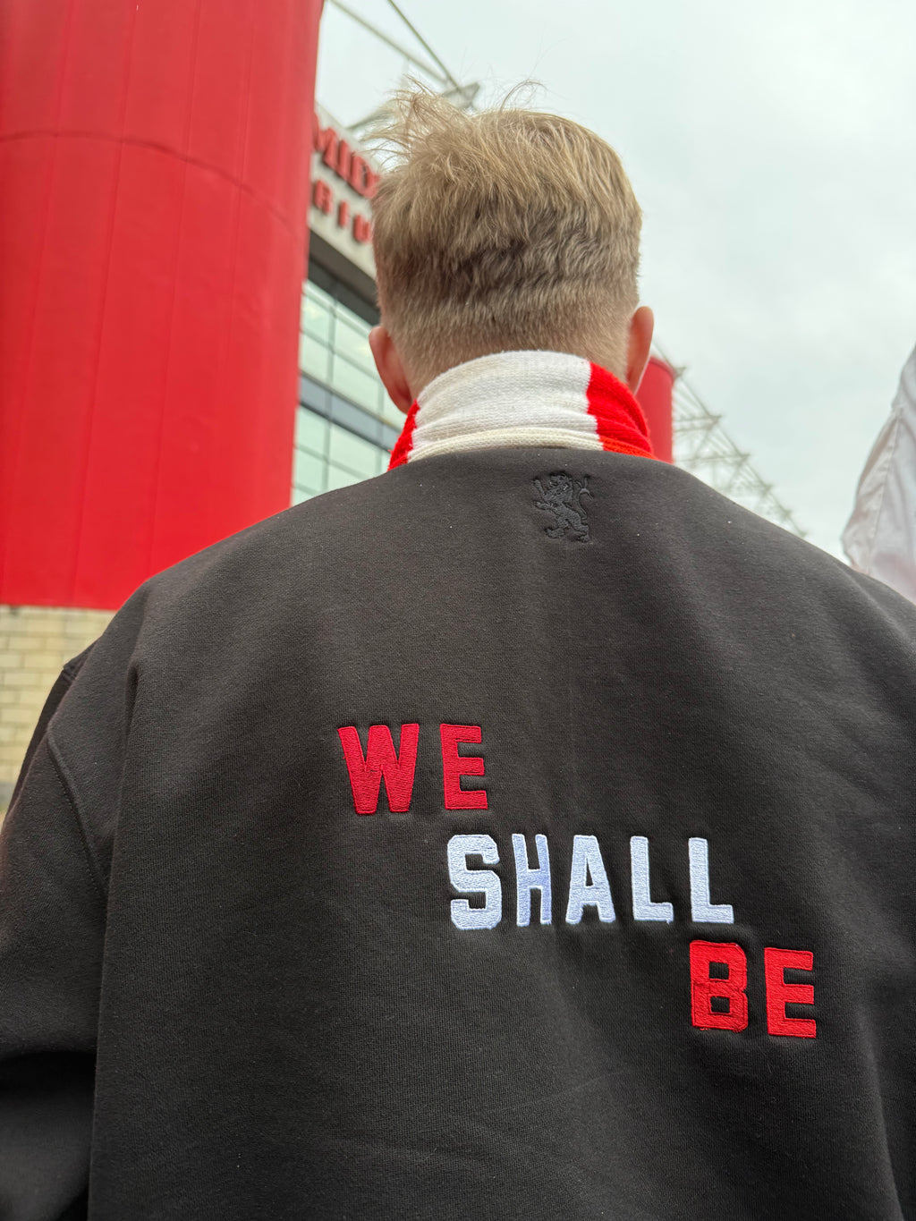 Person wearing a jacket with 'WE SHALL BE' text, standing in front of a red and white structure.