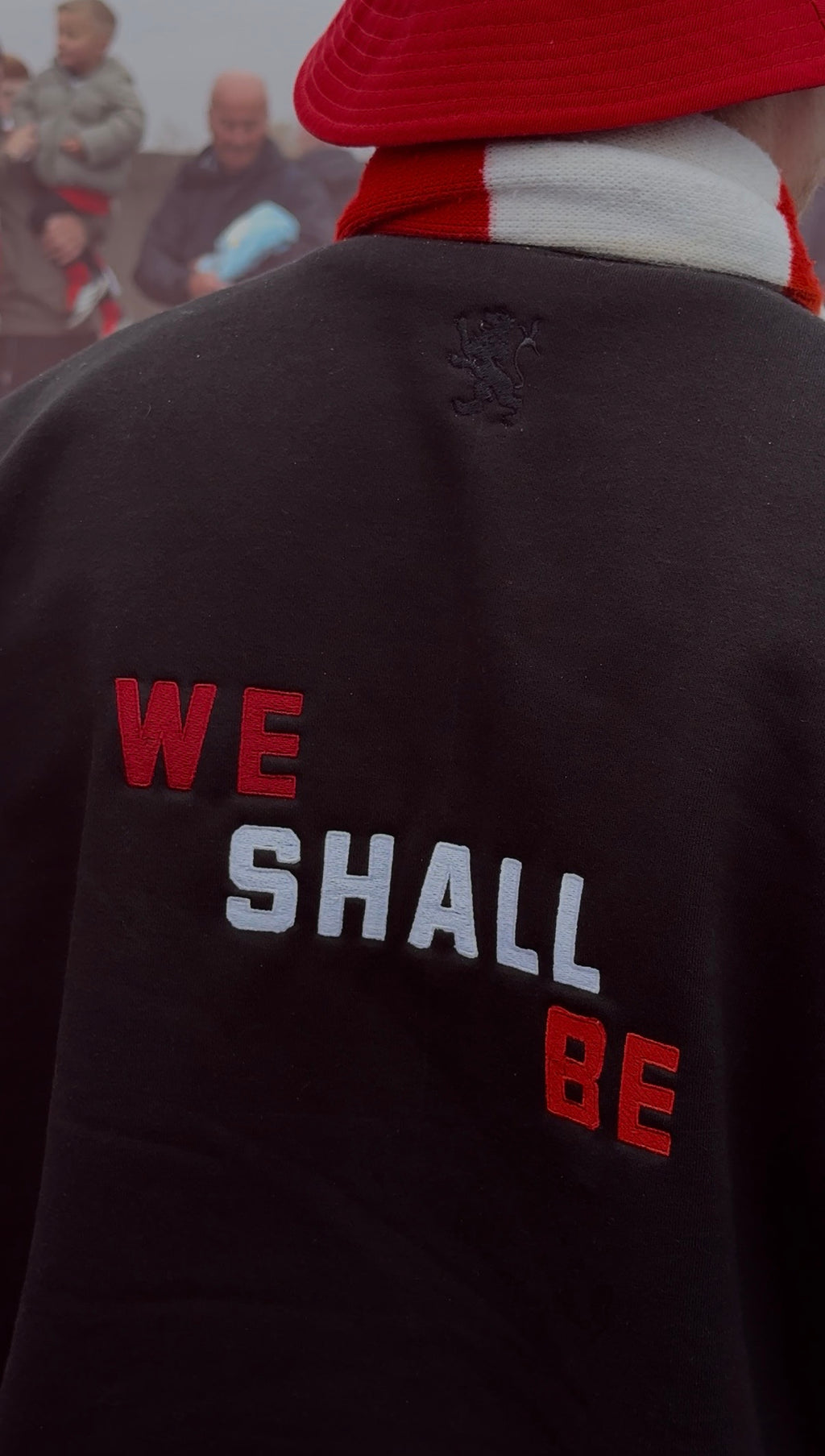 Person wearing a black jacket with 'WE SHALL BE' text, red cap, and white scarf.
