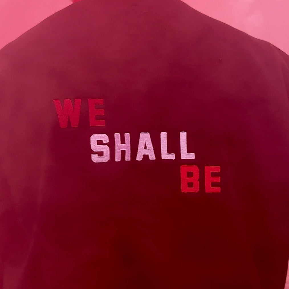 Person wearing a red bucket hat and black jacket with 'WE SHALL BE' text on a pink background