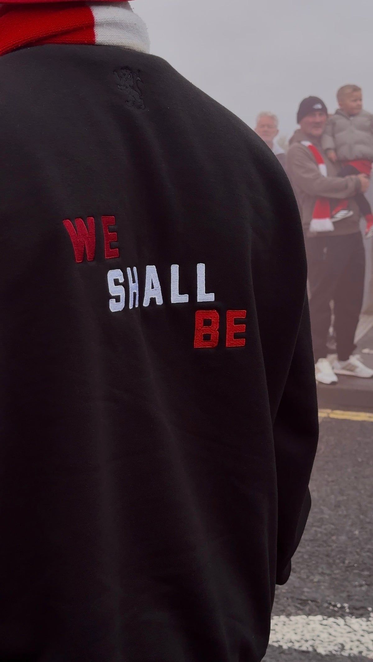 Person wearing a black jacket with 'WE SHALL BE' text, standing outdoors.